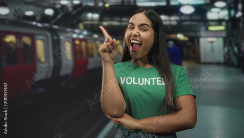 Tableau sur toile Woman pointing index finger up with volunteer shirt on a subway platform inside a building; community service confidence