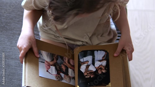 High angle view of a child sitting on the floor, holding and slowly opening a nostalgic photo album, remembering happy family moments and exploring her personal family history and roots