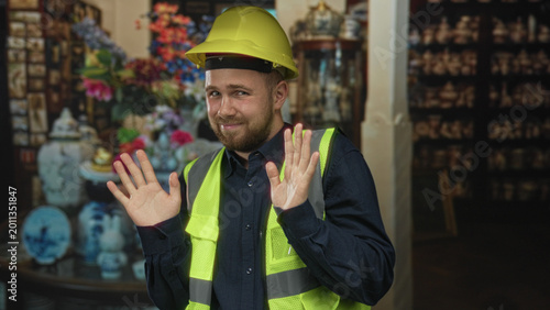 Obraz na plátně Man builder in hardhat and hi vis vest with hands raised palms out in building, gesturing a hesitant stop or surrender; embarrassment