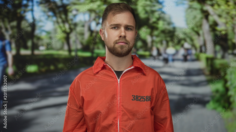 Αυτοκόλλητο Man in orange prison jumpsuit with number 382586 standing and looking at camera on a tree lined park path; solitude