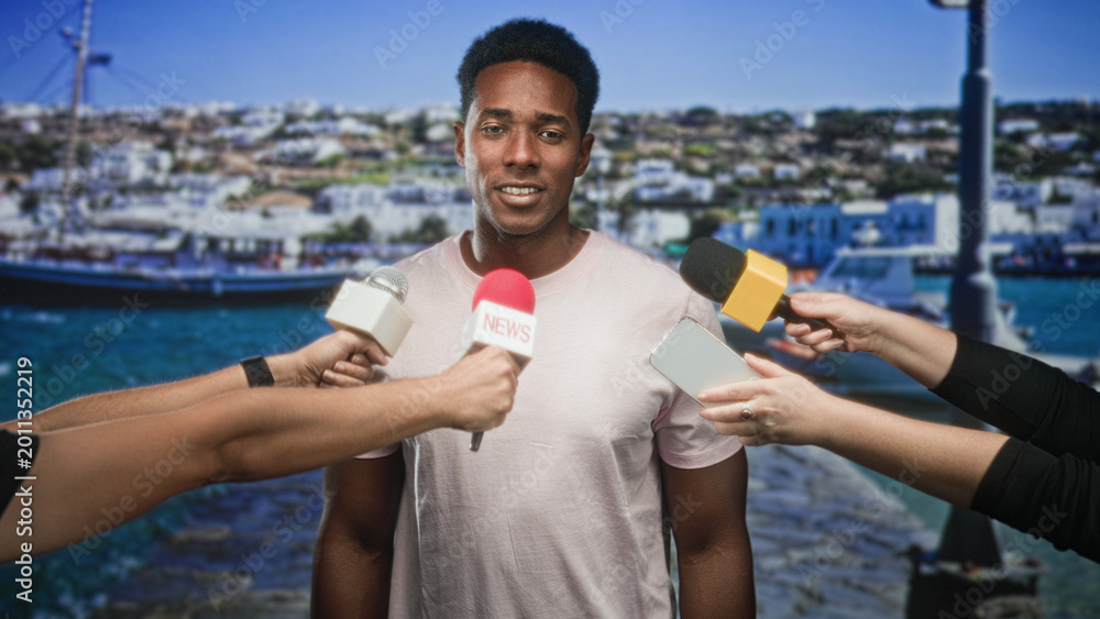 Naklejka premium Man answering questions to reporters with microphones and smartphone at harbor pier, boats and town behind him; confidence public statement.