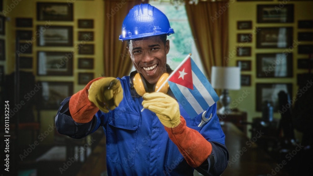 Fototapeta premium Man in hardhat holding cuban flag, wearing yellow gloves and earmuffs, smiling while pointing finger toward camera in building; national pride.