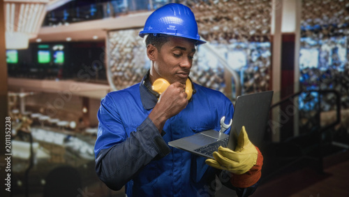 Fotografia Man in hardhat wearing blue coverall and noise cancel headphones holding laptop with wrench and hand to chin in building; focused problem solving