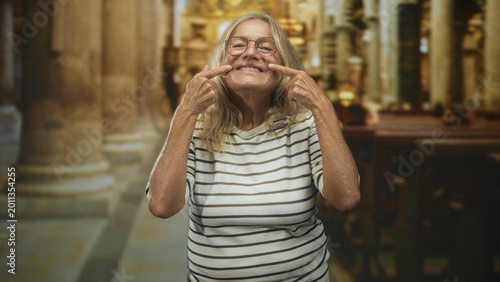 Foto Senior woman pointing fingers to her cheeks while smiling in a church nave among pews and stone columns; joy