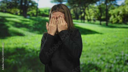 Photos Blonde young woman covering eyes with hands and wiping tears in a sunlit green park with trees and grass; private sadness