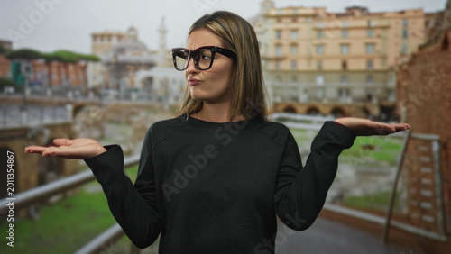 Cuadro en lienzo Woman blonde wearing glasses holds palms up at roman ruins building with puzzled expression and shrugged shoulders while standing on walkway; uncertainty
