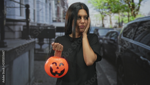 Obraz na płótnie Woman holds orange jackolantern bucket in left hand and shrugs with right palm up on a city street in front of parked cars; uncertainty waiting