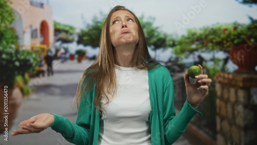 Obraz na płótnie Woman holding lime near face and grimacing with closed eyes and open mouth on street, wearing green cardigan and white shirt; disgust