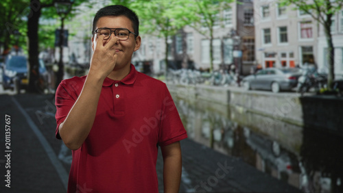 Tableau sur toile Man pinches nose on street canal path wearing red polo and glasses, eyes closed and subtle pointing gesture visible; disgust