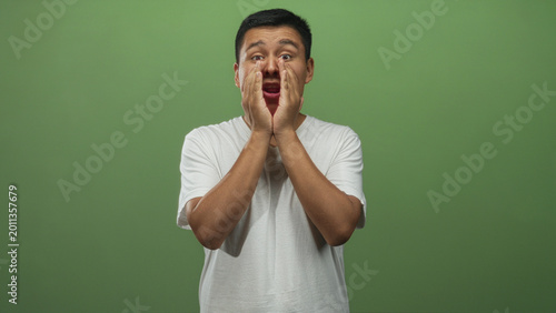 Foto Young hispanic man cupping hands to mouth shouting in green studio, facing forward with open mouth and intense expression; surprise