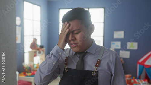 Foto Man teacher holds forehead for headache in kindergarten classroom building, removing glasses and rubbing temples; stress overload