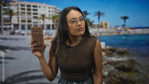 Photos Woman holds chocolate bar up in hand by oceanfront building on a sunny boardwalk, wearing brown sleeveless top, glasses and jeans; curiosity