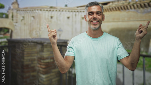 Photos Man pointing both index fingers then crossing arms, hands visible, standing by an old stone building with railing and lamp; curiosity reflection