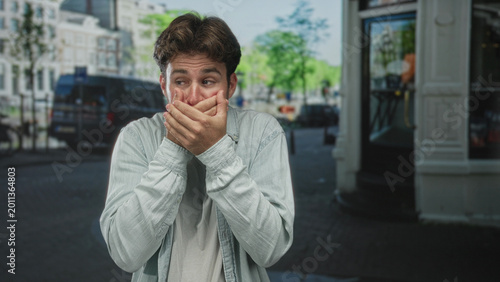 Obraz na plátně Young hispanic man covering mouth with both hands on a busy city street, eyes wide and palms visible; silence secrecy