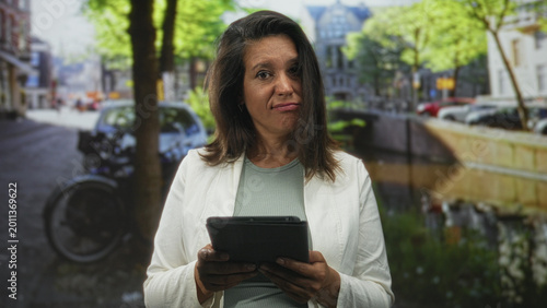 Εκτύπωση καμβά Woman holding tablet with both hands on a canal street, slight smirk and raised eyebrow, wearing white blazer; thoughtful focus