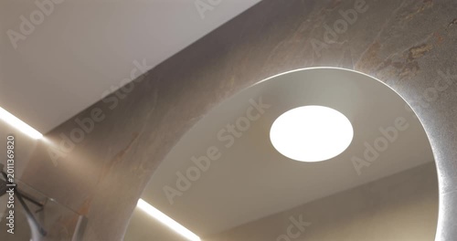 Ceiling light round mirror and wall faucet in smooth downward camera move, revealing light stone countertop with integrated sink and ribbed wood vanity as backlighting defines