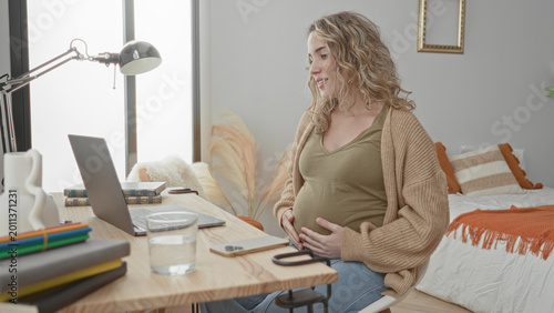 Φωτογραφία Pregnant woman holding belly at laptop in bedroom, smiling and cradling bump beside bed and desk while seated; contentment