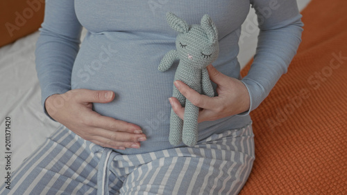 Canvas Print Woman holding a knitted bunny against her pregnant belly on a bed in a bedroom with hands cradling the bump; anticipation bonding