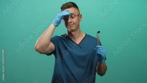 Foto Man dentist in blue scrubs holding dental mirror and instruments with gloved hand on forehead studio; tired patience