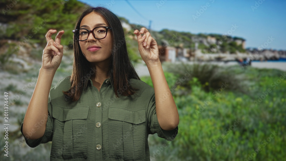 Fototapeta premium Young hispanic woman with glasses and brown hair making air quotes gesture in building near a green park and coastal dunes; playful skepticism.