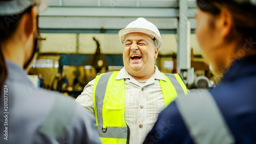 Fotografie Happy senior male engineer wearing a white hard hat and safety vest laughing loudly with his team during a break in a modern industrial factory setting