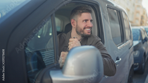 Foto Young man in driver's seat smiles while holding car keys and giving thumbs up through open window on a busy street; happiness trust