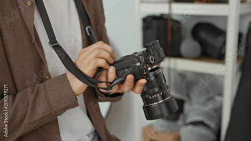 Cuadro en lienzo Young man adjusts camera dials while holding lens and strap in a photography studio with shelves of gear and softboxes visible; concentration craft