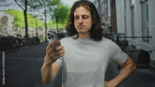 Fotografija Young hispanic man with long hair wearing grey t shirt holding smartphone in right hand with hand on hip on street looking at screen; annoyance distraction routine
