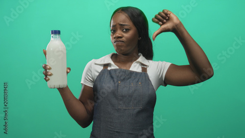 Valokuva Woman in apron holds milk bottle with thumb down and frowning expression, standing before a green studio backdrop; disgust