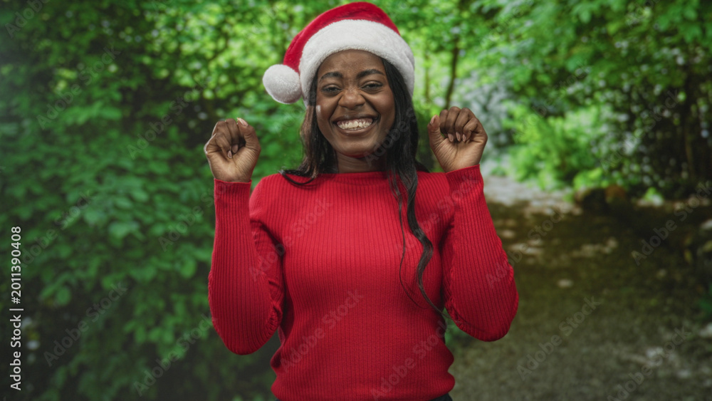 Fototapeta premium Woman in santa hat and red sweater makes claw hands gesture while smiling broadly in forest setting, facing camera; holiday celebration joy.