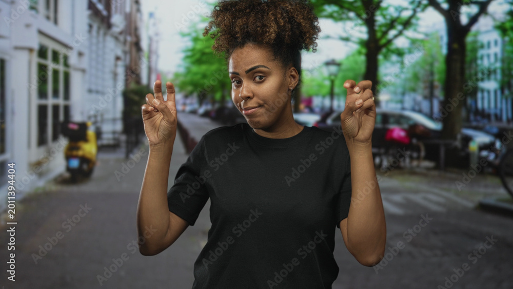 Fototapeta premium Woman making air quotes with fingers on a city street, hands raised, wearing black tee and curly hair, face expressed with mild smirk; sarcasm commentary.
