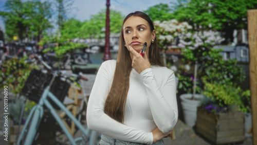 Canvastavla Young woman holding a vaporizer to her chin with finger on face, posed on a street near bicycles and planters; contemplation