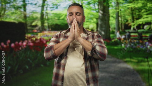Slika na platnu Man with palms together in prayer on a forest path, eyes closed, gray beard and bare hands visible; serenity reflection