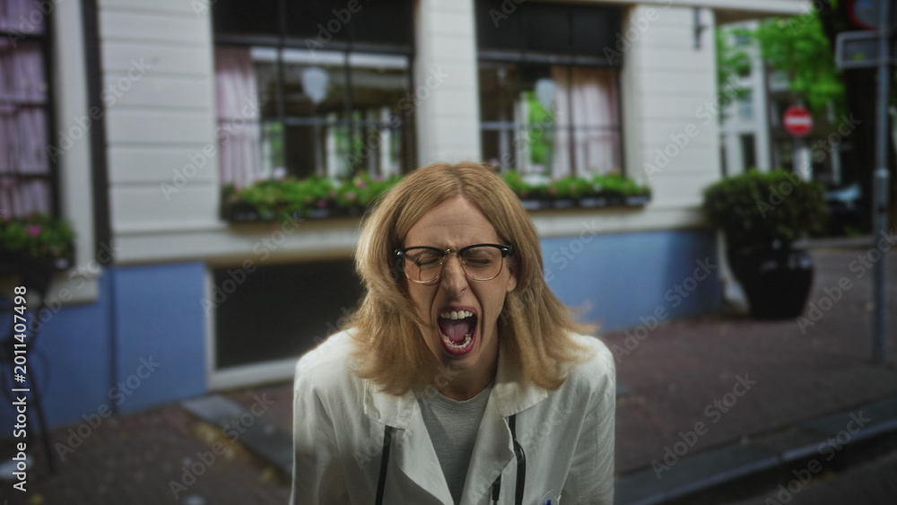 Fototapeta premium Woman doctor in lab coat and stethoscope shouting with mouth wide open and clenched hands on a city street outside a storefront building; stress urgency.