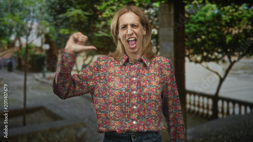 Foto Woman thumb pointing to chest while shouting with open mouth, floral shirt visible on street with trees and balustrade; confidence