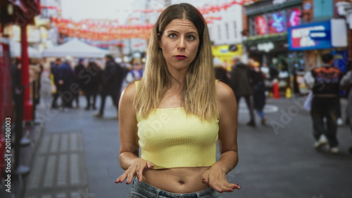 Young woman with bare midriff gesturing palms out while speaking on crowded street; concern communication.