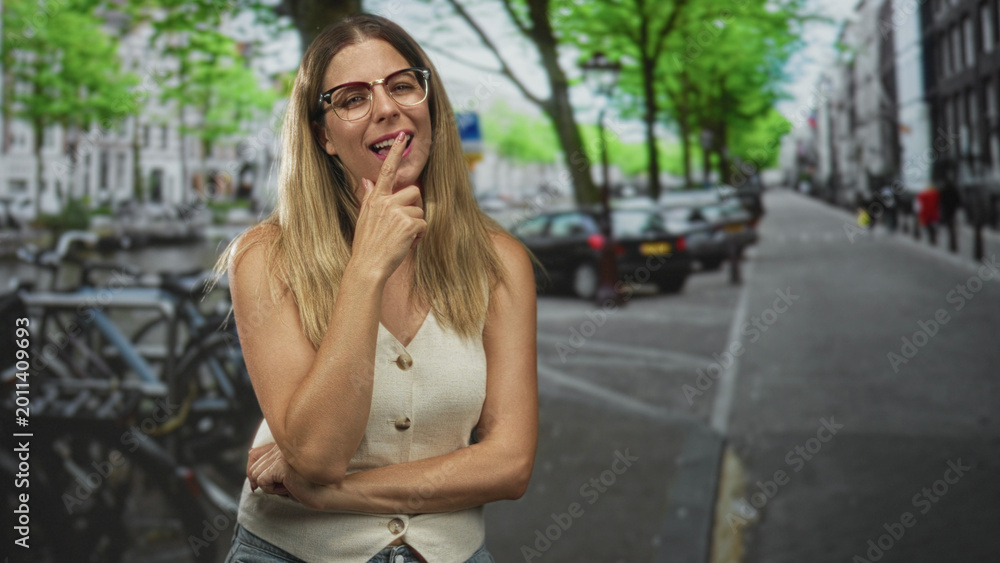 Fototapeta premium Woman with index finger to chin and glasses, smiling with arms crossed at waist on street near parked cars and bicycles; thoughtful curiosity.