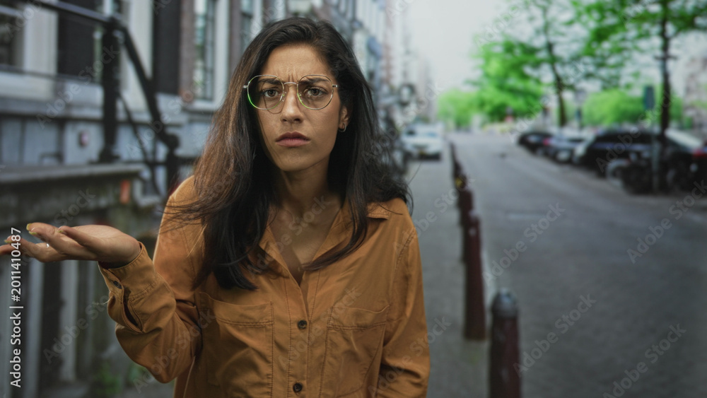 Fototapeta premium Woman shrugging with palms up on a city street wearing brown shirt and glasses, looking puzzled at an empty sidewalk; confusion questioning.