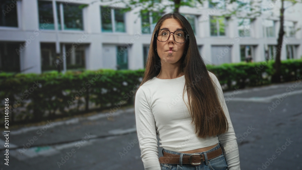 Fototapeta premium Woman wearing glasses smiles and turns head on a city street, long hair draped over shoulder, casual jeans and belt visible; confidence curiosity.