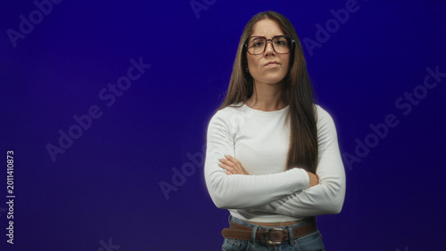 Bild auf Leinwand Woman with glasses, arms crossed, long hair, wearing white top, jeans and brown belt against a deep purple studio backdrop; quiet determination