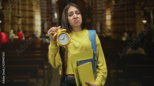 Принти на полотні Young female student in yellow sweater holds a yellow alarm clock and folders with a blue backpack, pouting while raising the clock in a building with pews; lateness anxiety
