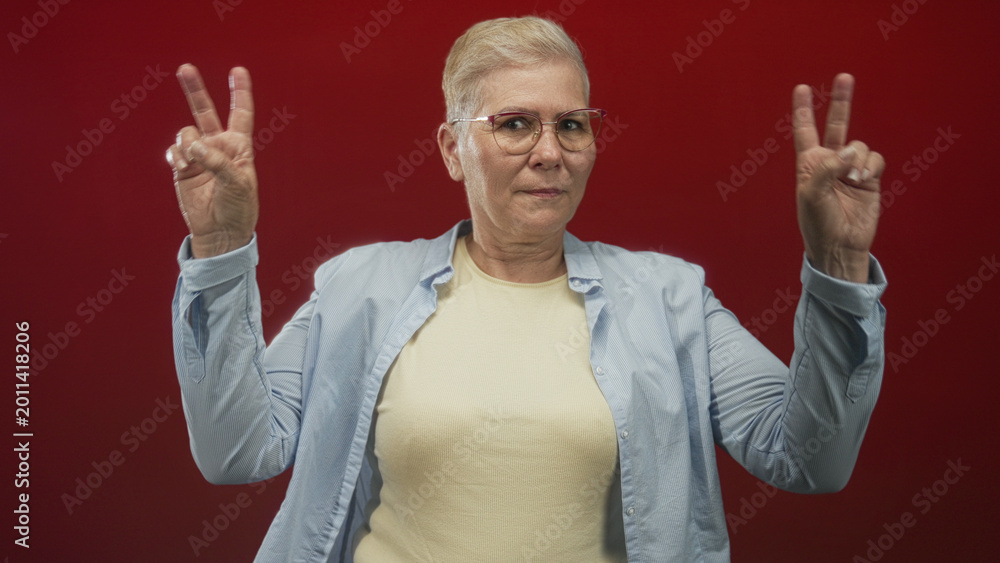 Fototapeta premium Woman making air quotes with both hands, wearing glasses and a light button shirt in a red studio; playful skepticism.