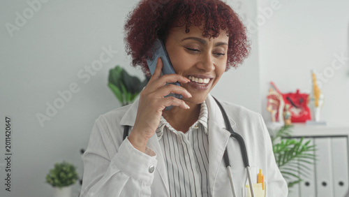 Woman doctor holding smartphone to ear and reading clipboard with stethoscope, smiling in building; professional care.
