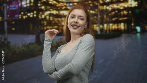 Canvas Print Young redhead woman touching her shoulder while flipping hair on illuminated city street at night; playful confidence