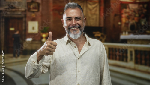 Fotografía Man with grey beard giving thumbs up inside an ornate catholic church building, smiling and looking at camera in a casual linen shirt; joy gratitude faith