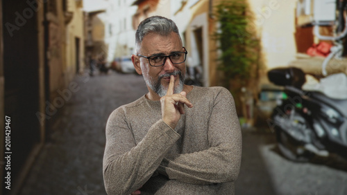 Photos Man with index finger to lips for silence on a narrow city street, wearing glasses and sweater, beard visible; playful secrecy