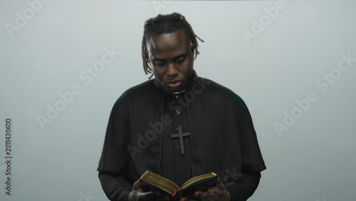 Εκτύπωση καμβά Young african american priest in black cassock reading a gilded book, holding it with hands in a pale gray studio; faith contemplation devotion