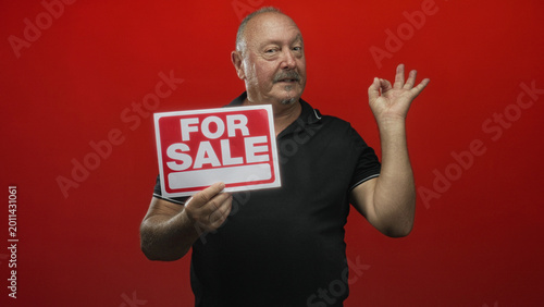 Foto Man holds a red for sale sign while making an ok sign with his right hand against a studio backdrop, modest smile and relaxed pose; confidence approval