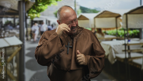 Photos Man monk in brown robe covers face with hand and wipes tears while standing by outdoor cafe stalls on street; sorrow reflection repentance