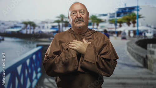 Tableau sur toile Man monk with hands pressed to chest wearing brown robe and cross, standing on street waterfront promenade in prayer; devotion reflection solitude
